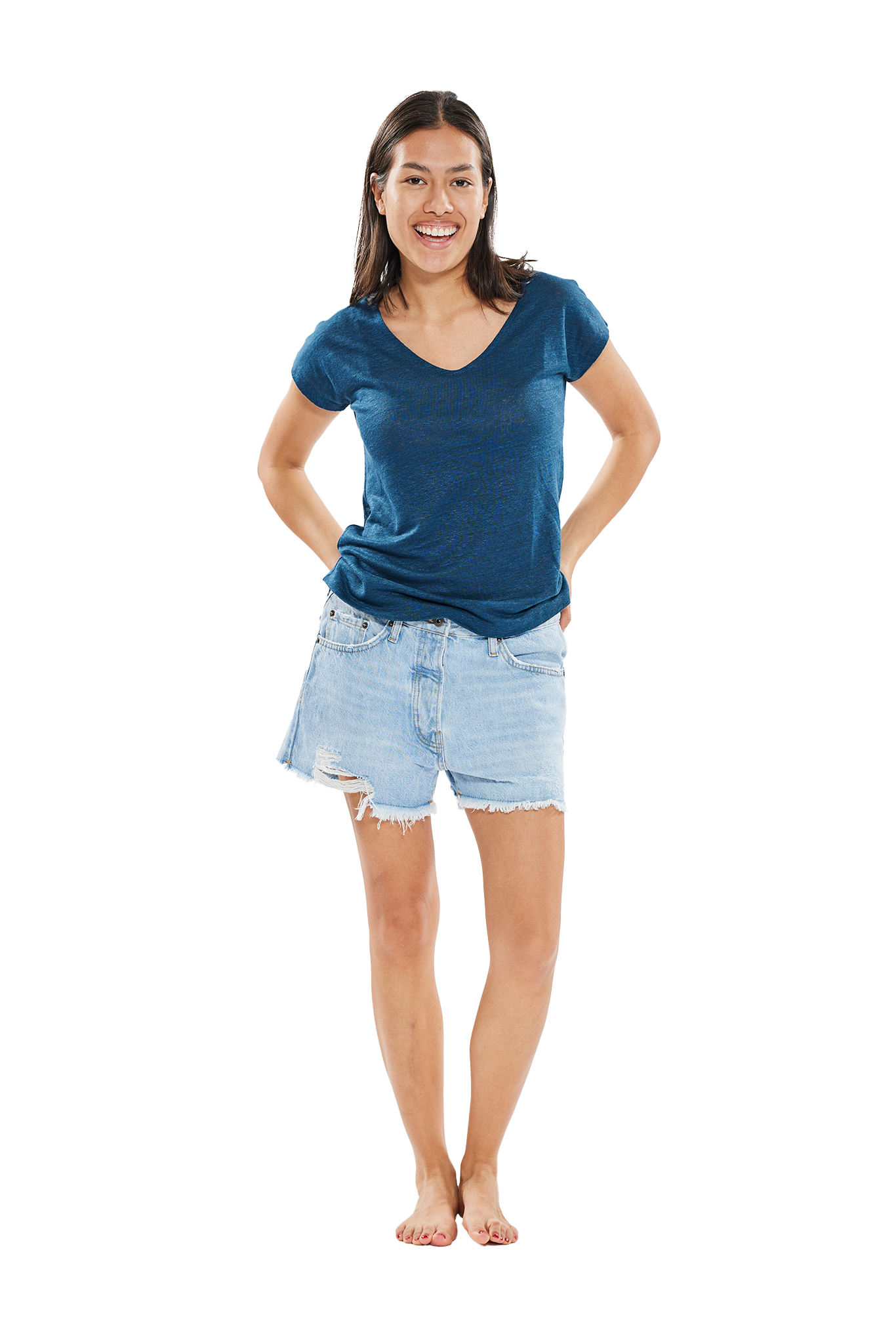 Lächelnde Frau in blauem T-Shirt und ausgefransten Jeansshorts, barfuß stehend vor einem weißen Hintergrund.