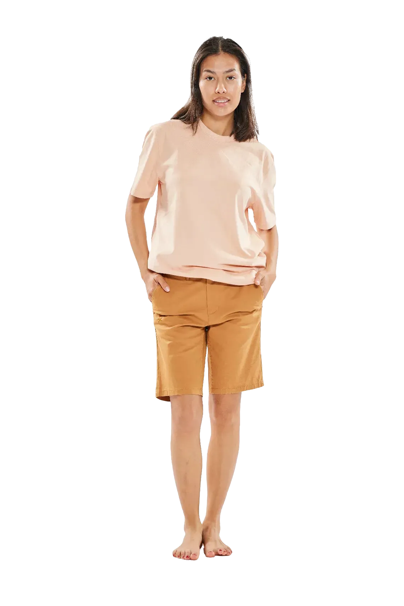 Junge Frau mit schulterlangem Haar, die ein helles T-Shirt und braune Bermuda-Shorts trägt, steht barfuß mit den Händen in den Taschen vor einem weißen Hintergrund.