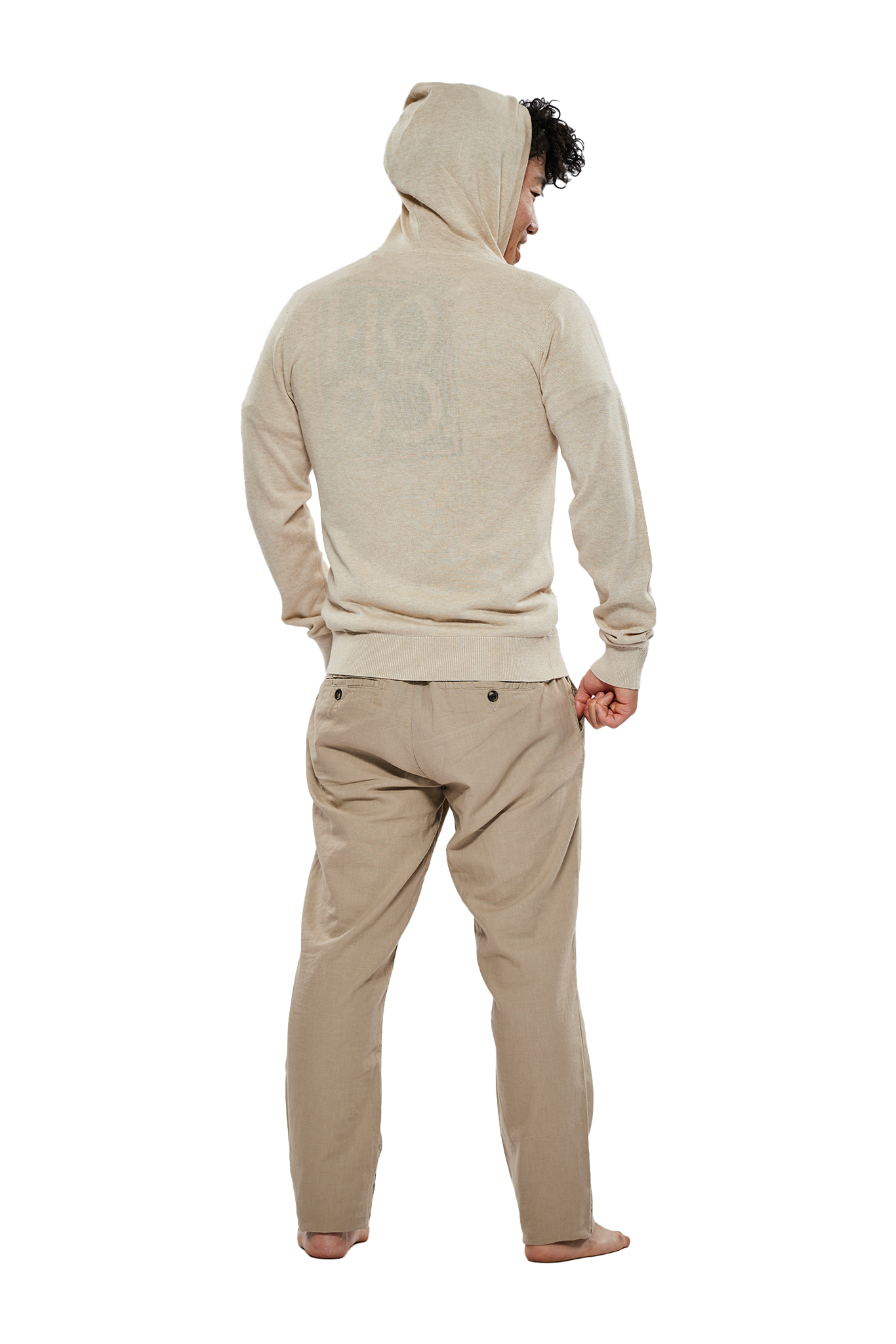 Ein Mann mit lockigem Haar steht barfuß mit dem Rücken zur Kamera vor einem weißen Hintergrund. Er trägt eine beige Strickjacke mit Kapuze, die über den Kopf gezogen ist, und beige Hosen.