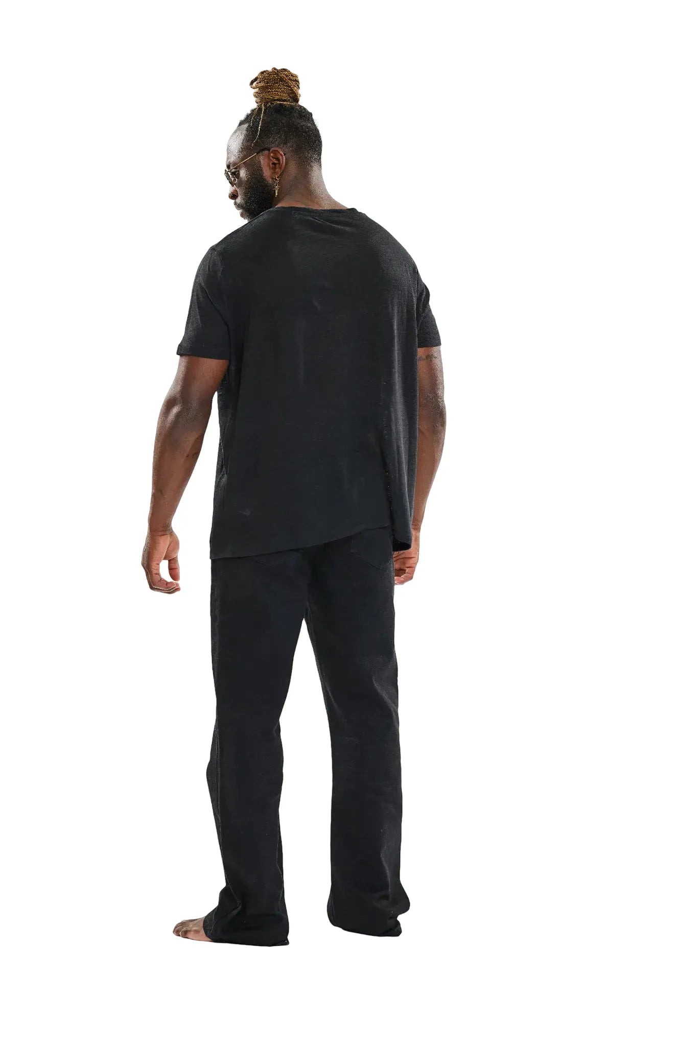 Rückansicht eines Mannes mit Dreadlocks, gekleidet in ein schwarzes T-Shirt und schwarze Hose, barfuß stehend vor einem weißen Hintergrund.