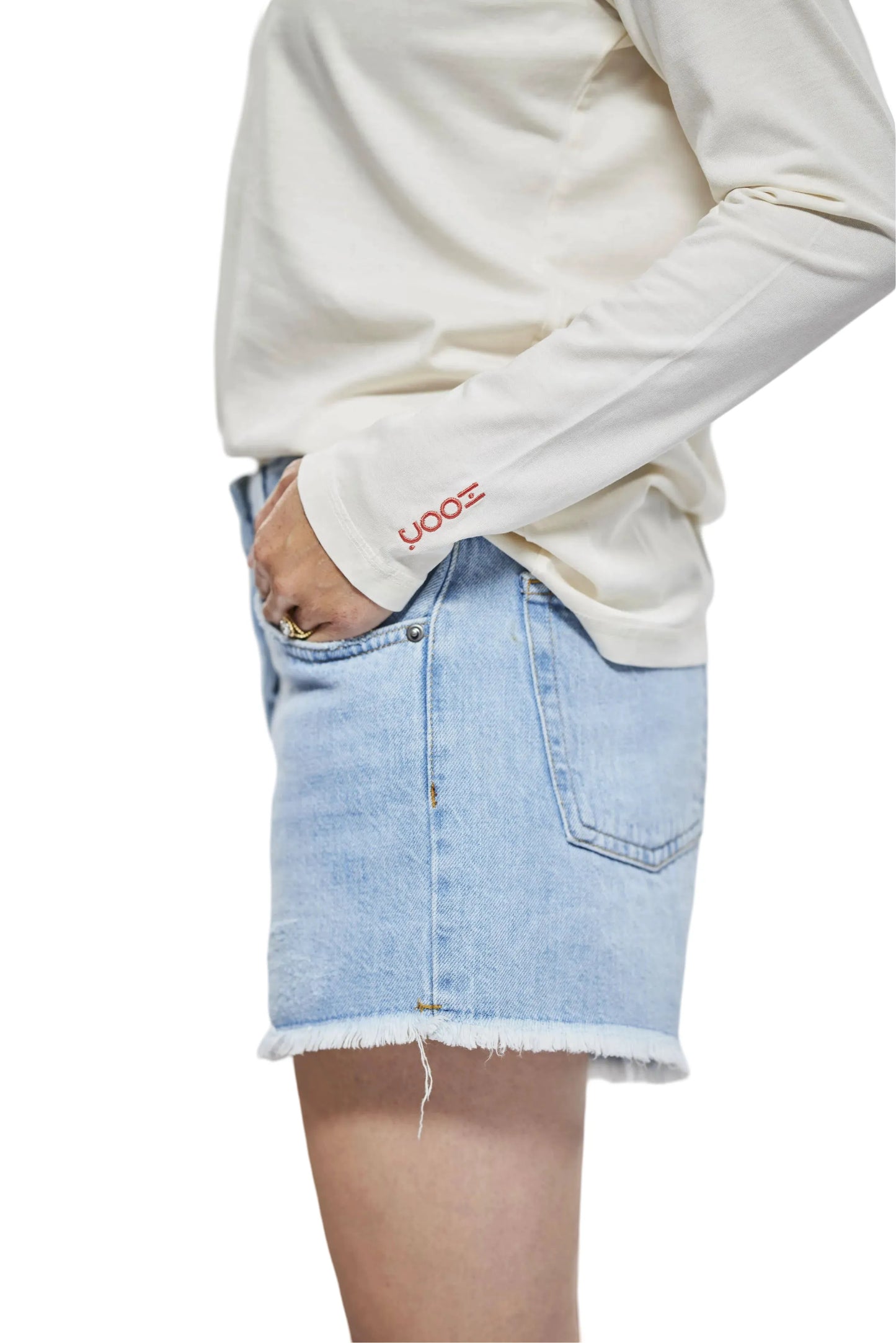 Nahaufnahme der rechten Seite einer Frau in einem weißen Langarmshirt und hellblauen Jeansshorts. Auf dem Ärmel des Shirts ist ein kleines rotes Logo zu sehen. Die Frau hat eine Hand in der Hosentasche und steht vor einem weißen Hintergrund.