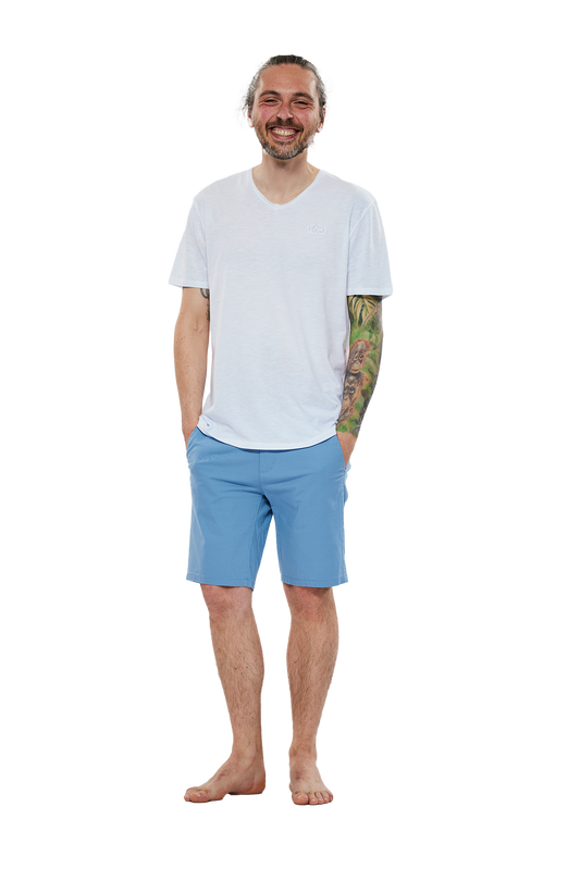 Lächelnder Mann in weißem T-Shirt und blauen Shorts, barfuß stehend vor einem weißen Hintergrund. Sein rechter Arm ist tätowiert.