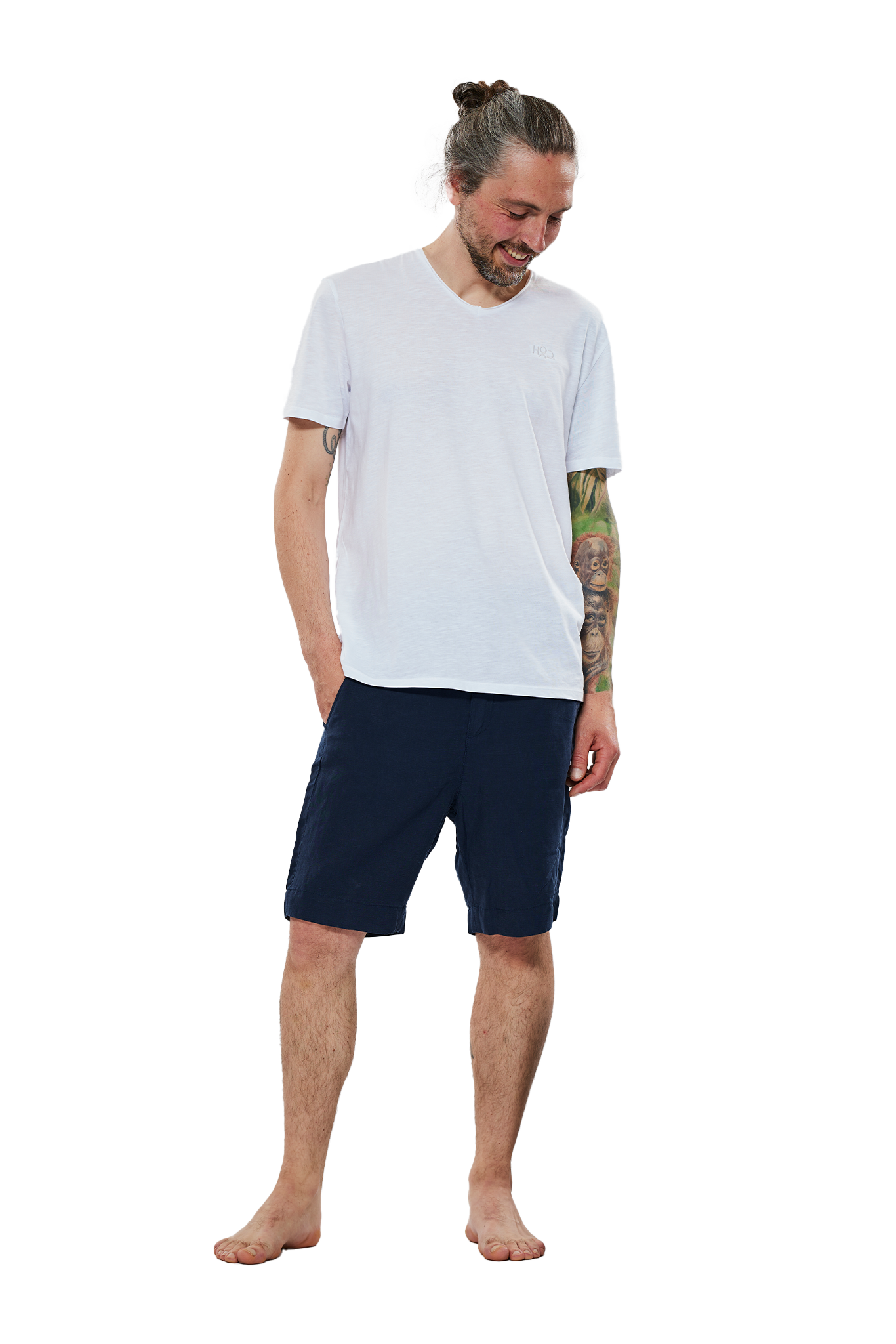 Lächelnder Mann mit Duttfrisur, tätowiertem Arm und weißem T-Shirt, der eine dunkelblaue Bermuda-Shorts trägt, steht barfuß vor einem weißen Hintergrund.