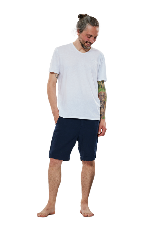 Lächelnder Mann mit Duttfrisur, tätowiertem Arm und weißem T-Shirt, der eine dunkelblaue Bermuda-Shorts trägt, steht barfuß vor einem weißen Hintergrund.