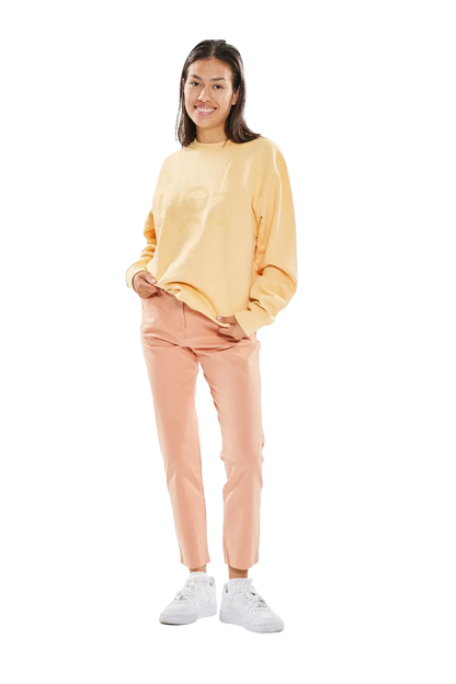 Eine junge Frau mit schulterlangem, dunklem Haar trägt einen gelben Pullover und rosa Chino-Hosen. Sie steht in weißen Sneakers auf einem weißen Hintergrund und lächelt in die Kamera.