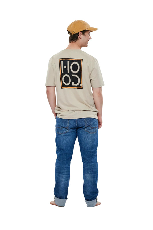 Ein junger Mann steht barfuß mit dem Rücken zur Kamera vor einem weißen Hintergrund. Er trägt ein beigefarbenes T-Shirt mit einem großen "HOOD"-Logo auf dem Rücken, dunkelblaue Jeans und eine beige Baseballkappe. Der Mann blickt zur Seite und lächelt leicht.