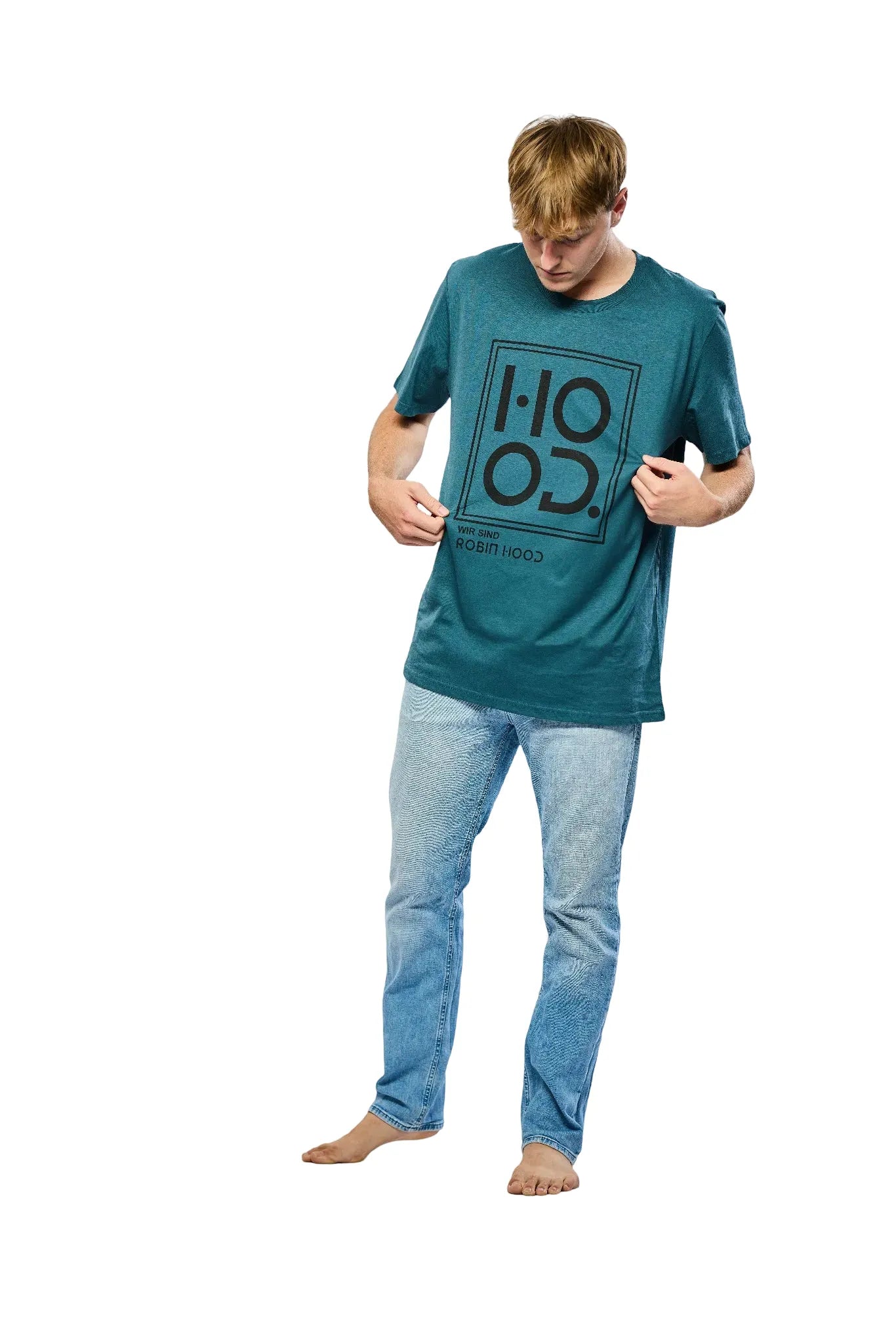 Mann in einem dunkelgrünen T-Shirt mit schwarzem grafischem Aufdruck und blauen Jeans, barfuß stehend und nach unten schauend vor einem weißen Hintergrund.