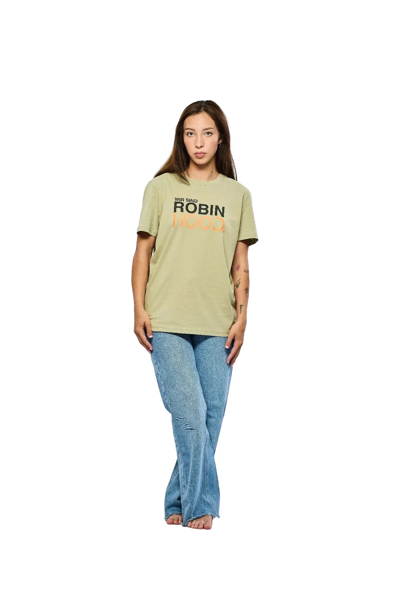 Eine junge Frau mit langen, glatten Haaren steht barfuß vor einem weißen Hintergrund. Sie trägt ein olivgrünes T-Shirt mit der Aufschrift "Wir sind ROBIN HOOD" in schwarzem und orangefarbenem Text sowie hellblaue, gemusterte Jeans. Die Frau schaut direkt in die Kamera, mit den Händen locker an den Seiten.