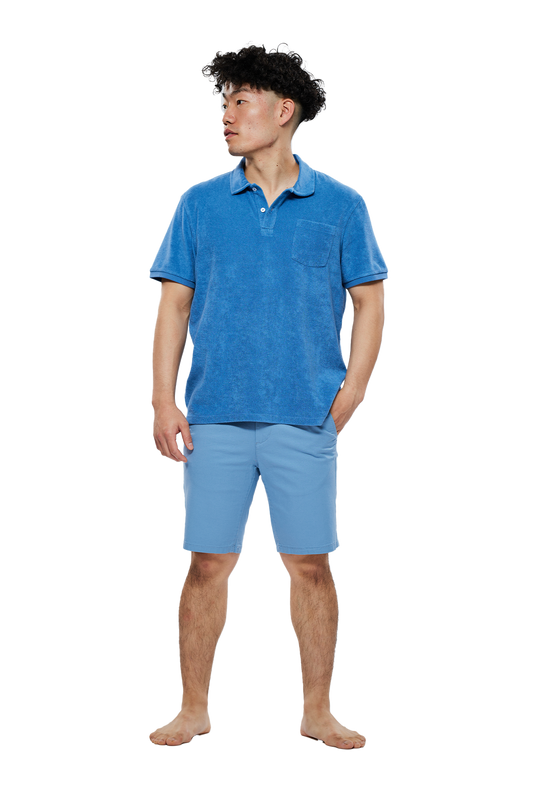 Ein junger Mann mit lockigem, dunklem Haar trägt ein blaues Frottee-Poloshirt und hellblaue Shorts. Er steht barfuß auf einem weißen Hintergrund und blickt nach links.