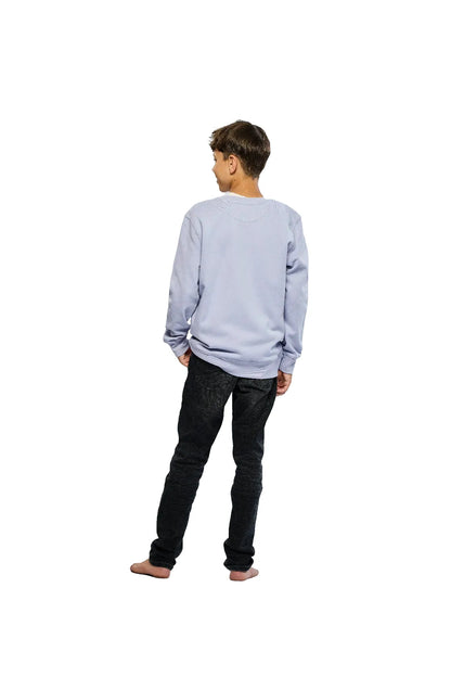 Rückansicht eines Jungen in einem lavendelfarbenen Pullover und dunklen Jeans, barfuß stehend und den Kopf zur Seite drehend, vor einem weißen Hintergrund.